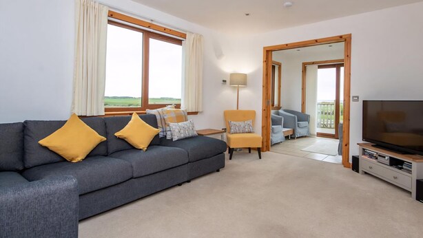 A modern living room with a grey sofa, yellow cushions, a wooden chair, a TV, and a large window offering a view of the outdoors. - 3 bedroom accommodation in Dornoch (Dornoch)