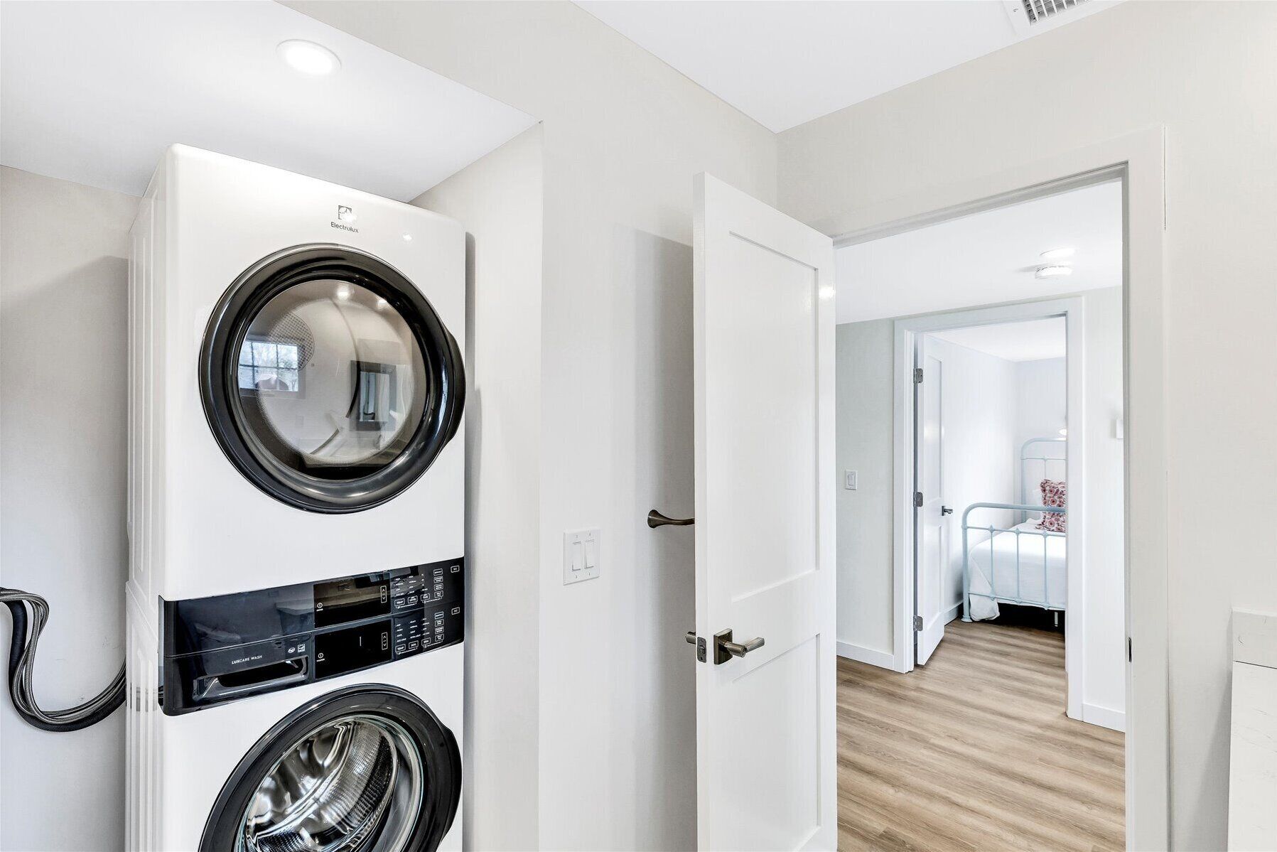 A laundry room with a front-loading washing machine and dryer, a door leading to a bedroom with a bed and a nightstand.
