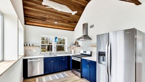 Une cuisine moderne dotée d’un réfrigérateur en acier inoxydable, d’un four encastré et d’une cuisinière à gaz. On y trouve des armoires bleues, un plafond en bois et une fenêtre donnant sur l’extérieur.