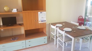 A dining area with a wooden table set for four, a TV on a stand, and a cabinet with a poster. - Trilocale Ulivo Baia Verde piano terra (Gallipoli)