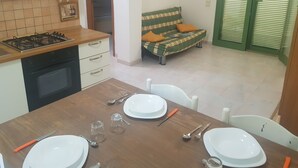 A dining area with a wooden table set for four, a kitchen with a gas stove and oven, a sofa with orange cushions, and a green door. - Trilocale Ulivo Baia Verde piano terra (Gallipoli)
