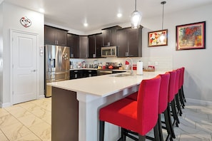 A modern kitchen with dark cabinets, stainless steel appliances, a white island with red bar stools, and artwork on the walls. - 8BR Near Disney•6 Kings•Heated Pool•Star Wars (Davenport)
