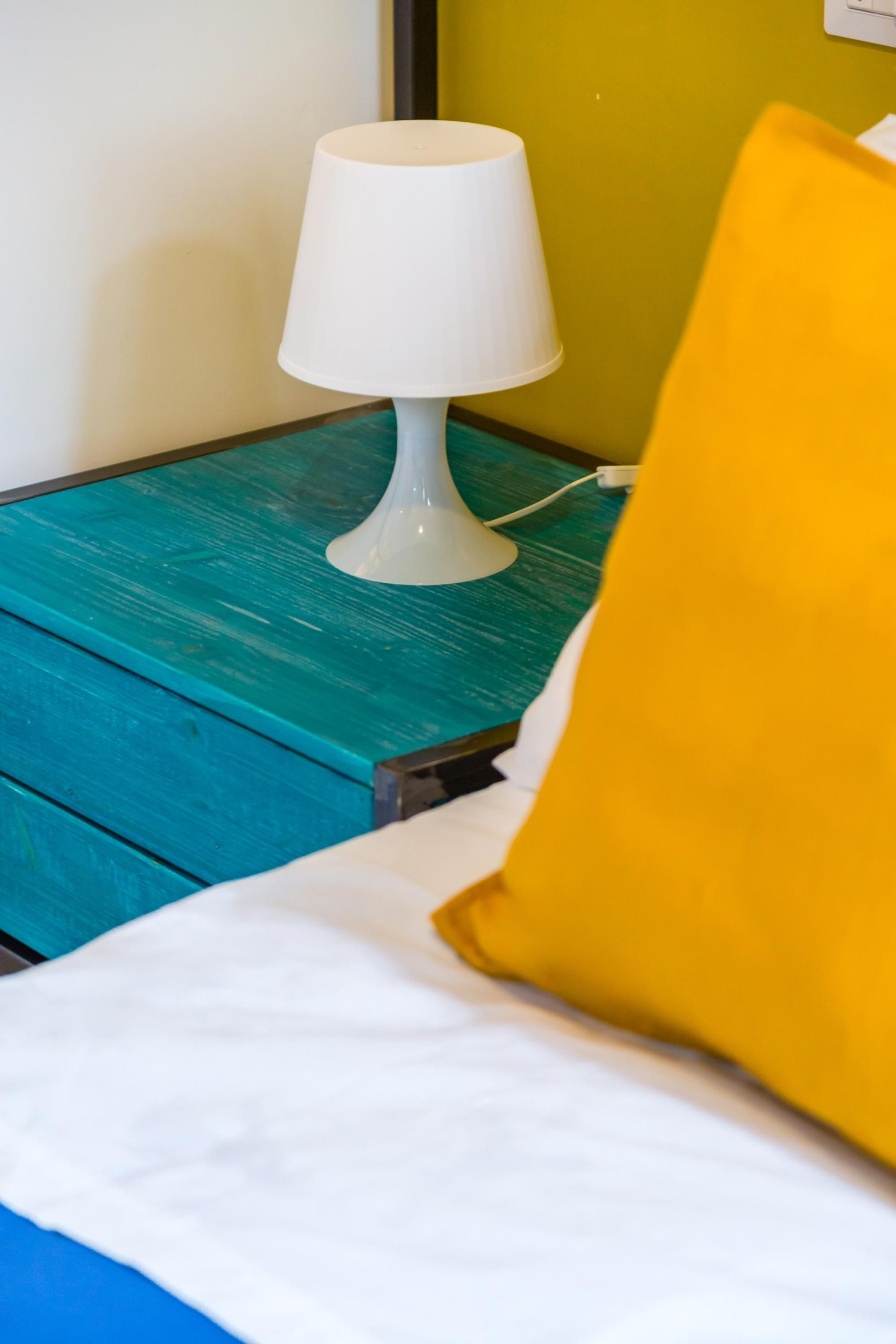 A bed with white bedding and a yellow pillow, a white lamp on a teal nightstand, and a green wall.
