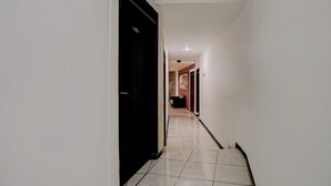Un pasillo estrecho con paredes blancas y suelo de baldosas. Hay puertas a ambos lados, una de ellas negra. El pasillo conduce a una habitación con una puerta visible al final.
