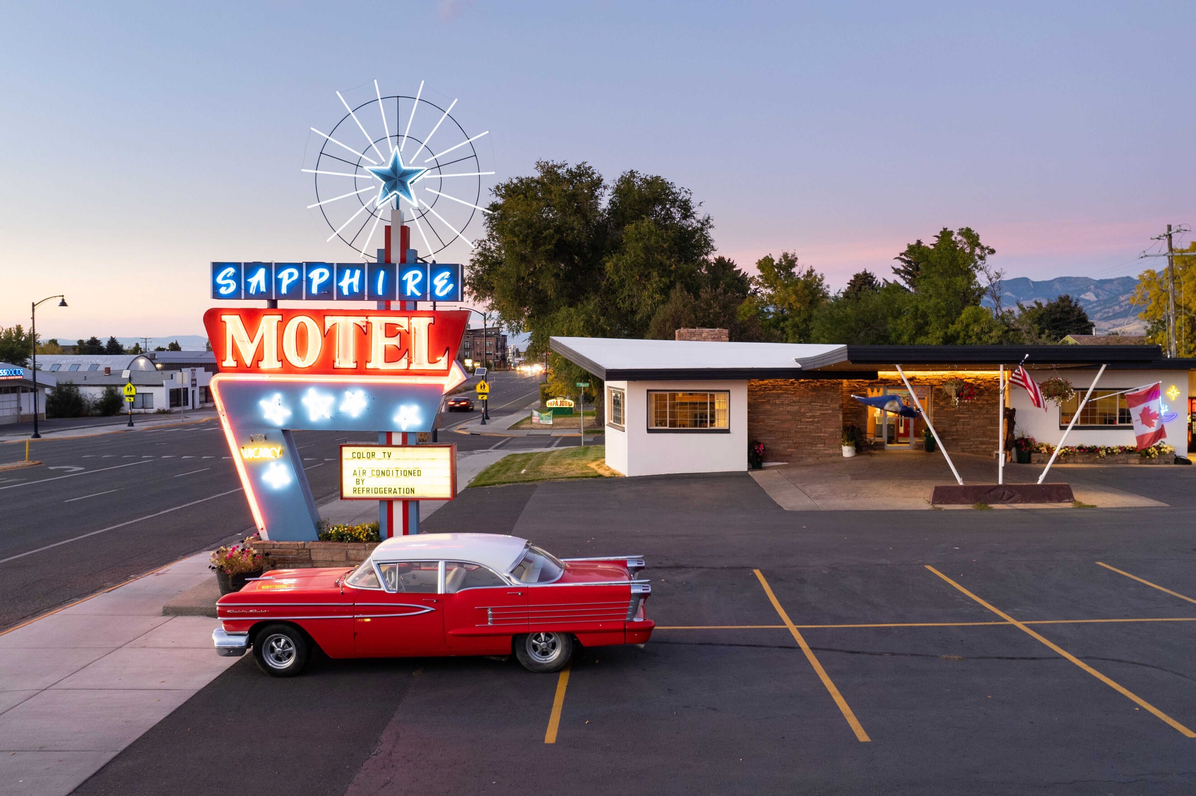 Et gammelt motel-skilt med et stjernenhjul og neonlys, en rød bil parkeret foran, og en mindre bygning med en hvid facade.