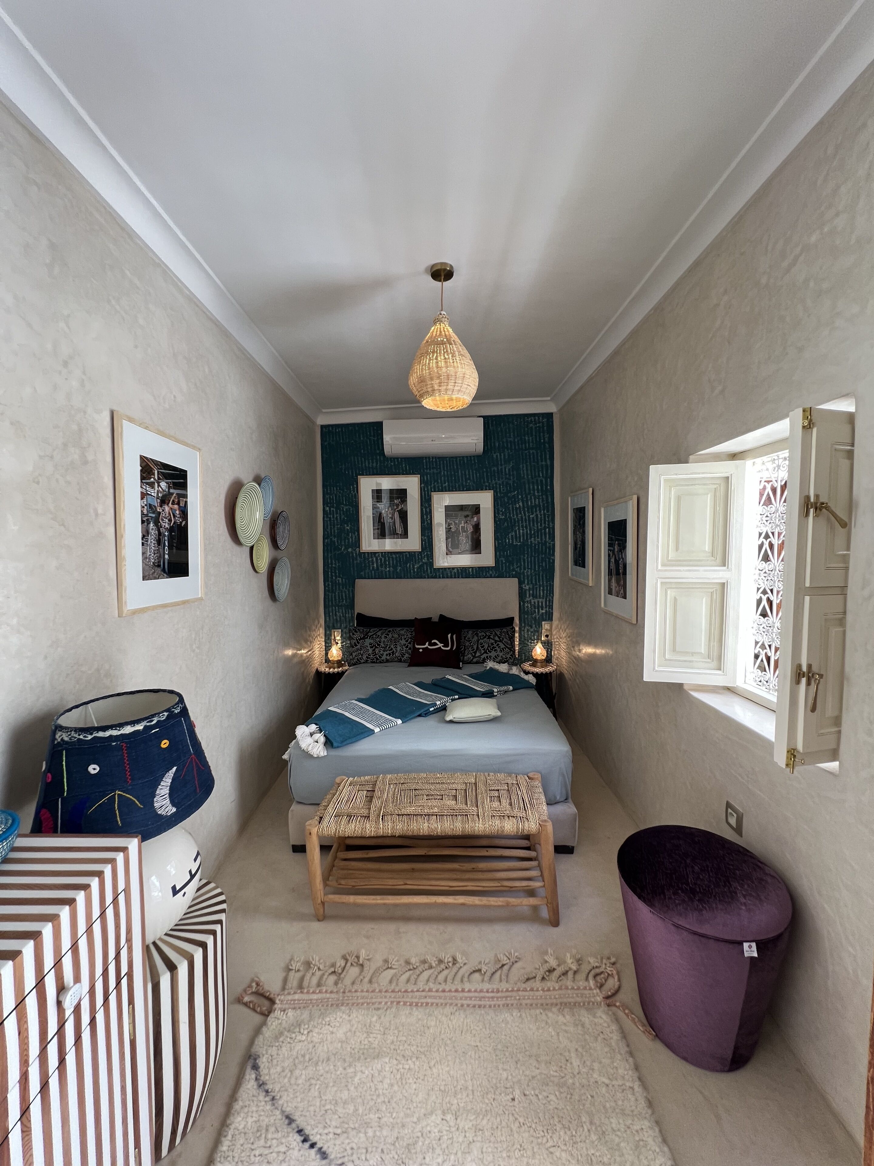 Ein Schlafzimmer mit Bett, Nachttischen, einer Hängelampe, einem Fenster mit verzierten Rollläden und einer Wand-Klimaanlage.