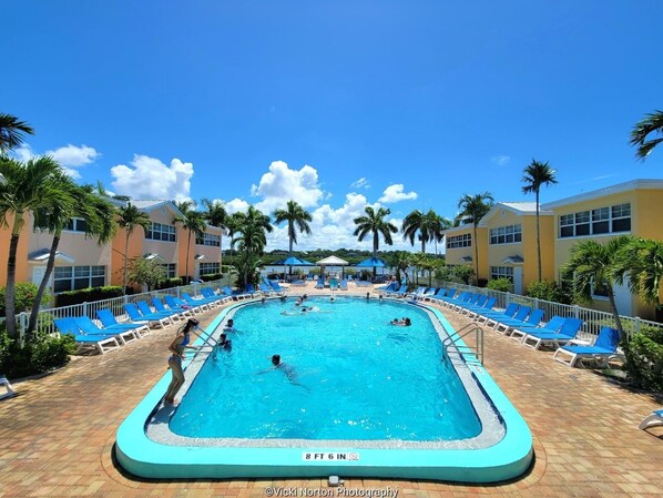 A swimming pool surrounded by blue lounge chairs and palm trees, with a clear blue sky above.