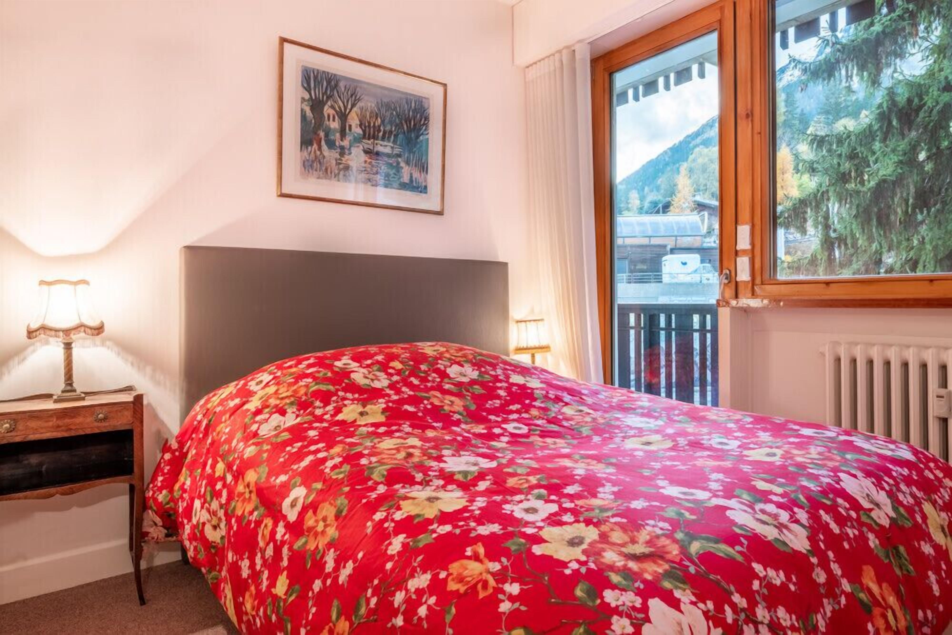 Un dormitorio con una cama grande, una mesita de noche con lámpara, un cuadro en la pared y una ventana con vistas a árboles y montañas.