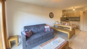 A living room with a grey sofa, a wooden coffee table, and a kitchen area with wooden cabinets and a microwave.