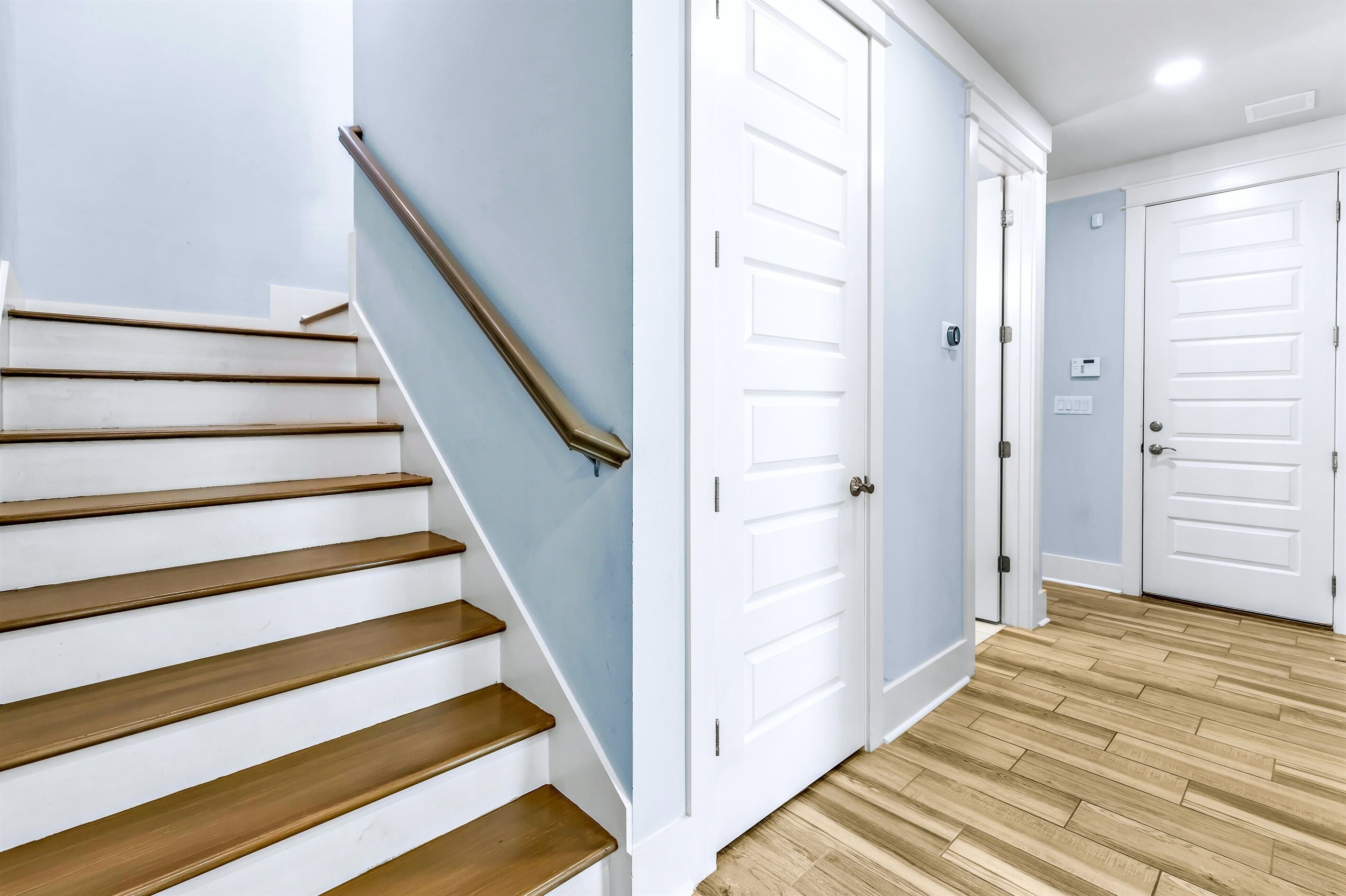 Un escalier avec des marches en bois et une rampe métallique, menant à un couloir avec des portes blanches et un mur bleu clair.