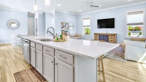 A modern kitchen with a large island, white cabinetry, and stainless steel appliances. There is a dining area with a white table and chairs, and a flat-screen TV mounted on the wall. - Sea Glass: Free golf cart, 2min to beach, Heated pool steps away, Bunk rooms (Miramar Beach)