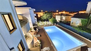 Une piscine avec des chaises longues, un escalier et une vue sur les maisons voisines la nuit.