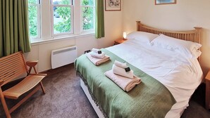 Una cama bien hecha con una manta verde y toallas dobladas, una silla de madera y una ventana con cortinas.