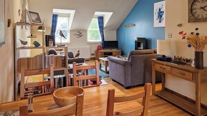 Una sala de estar acogedora con una mesa de comedor de madera, sillas, un sofá y un televisor. Hay estanterías y una pequeña zona de cocina.