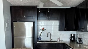 Une cuisine moderne avec des meubles de couleur sombre, un réfrigérateur en acier inoxydable, une table de salle à manger ronde en bois et des chaises en osier, ainsi qu’un ventilateur de plafond.