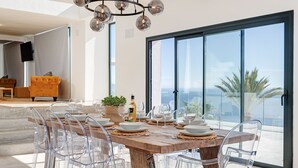A dining area with a large table, clear chairs, a chandelier, and a view of the ocean through large windows. - Villa Sofia - Four Bedroom Villa, Sleeps 10 (Salobreña)