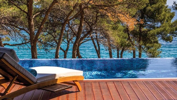 An infinity pool with a wooden deck and lounge chair, surrounded by trees and overlooking the sea.