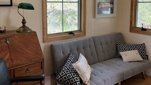 A living room with a grey sofa, a wooden cabinet, a green desk lamp, and two windows with views of trees. - Cozy secluded cabin on a working cattle ranch (Clifton)