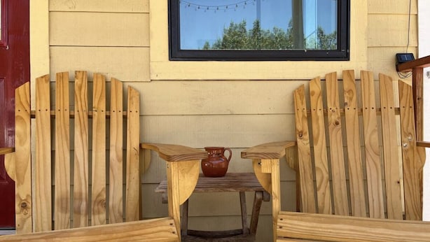 Two wooden Adirondack chairs with a small table between them, a window with a view of trees, and a red door. - Cozy secluded cabin on a working cattle ranch (Clifton)