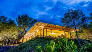 Un bâtiment moderne doté d’un toit en pente et de grandes fenêtres en verre, entouré d’arbres et d’un jardin aménagé.
