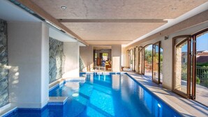 A modern indoor swimming pool with clear blue water, stone walls, and large sliding glass doors leading to an outdoor area.