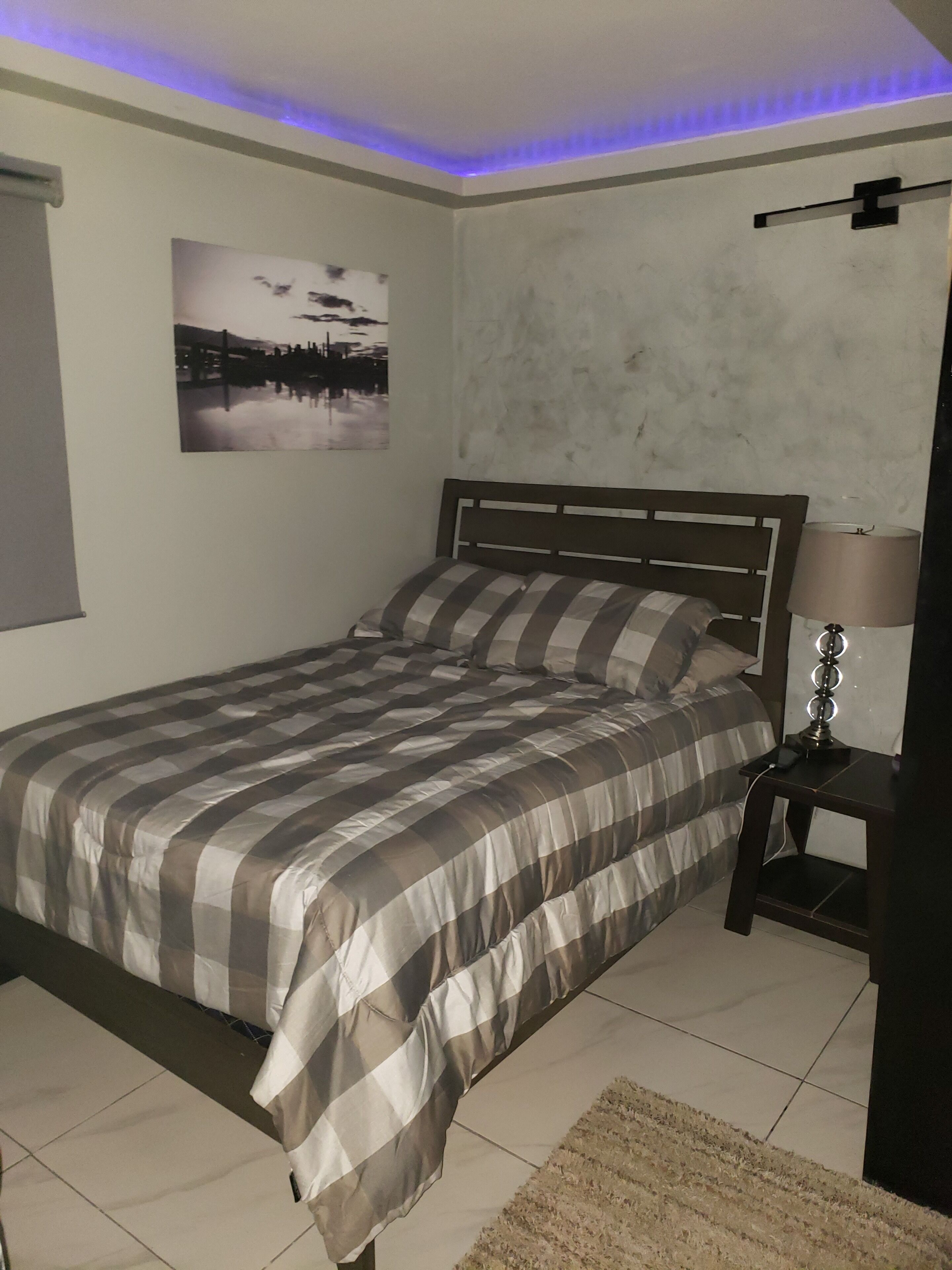 A bedroom with a bed, a bedside table with a lamp, a picture on the wall, and a ceiling light.