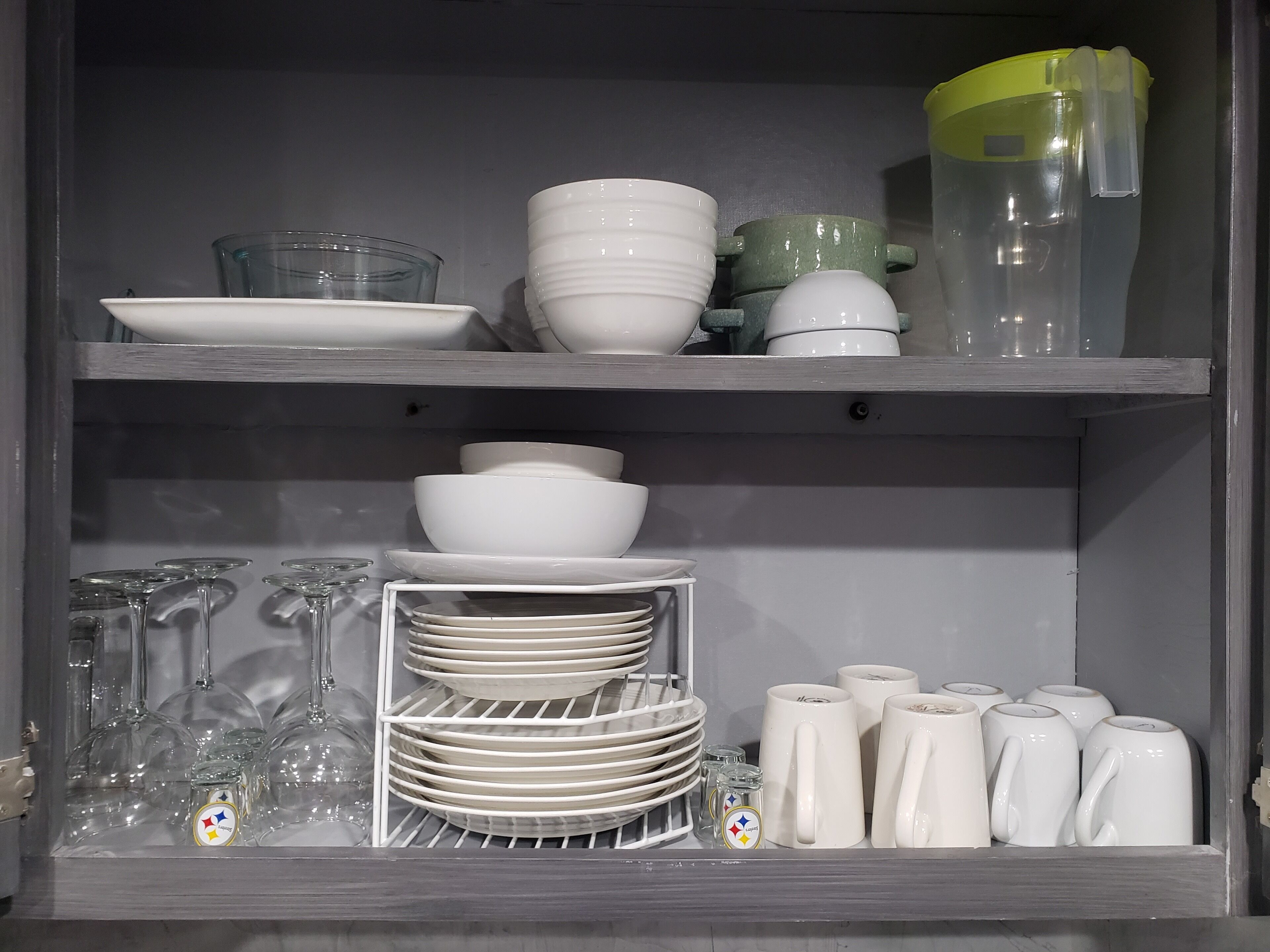 A kitchen cabinet with shelves holding various white dishes, glassware, and mugs.