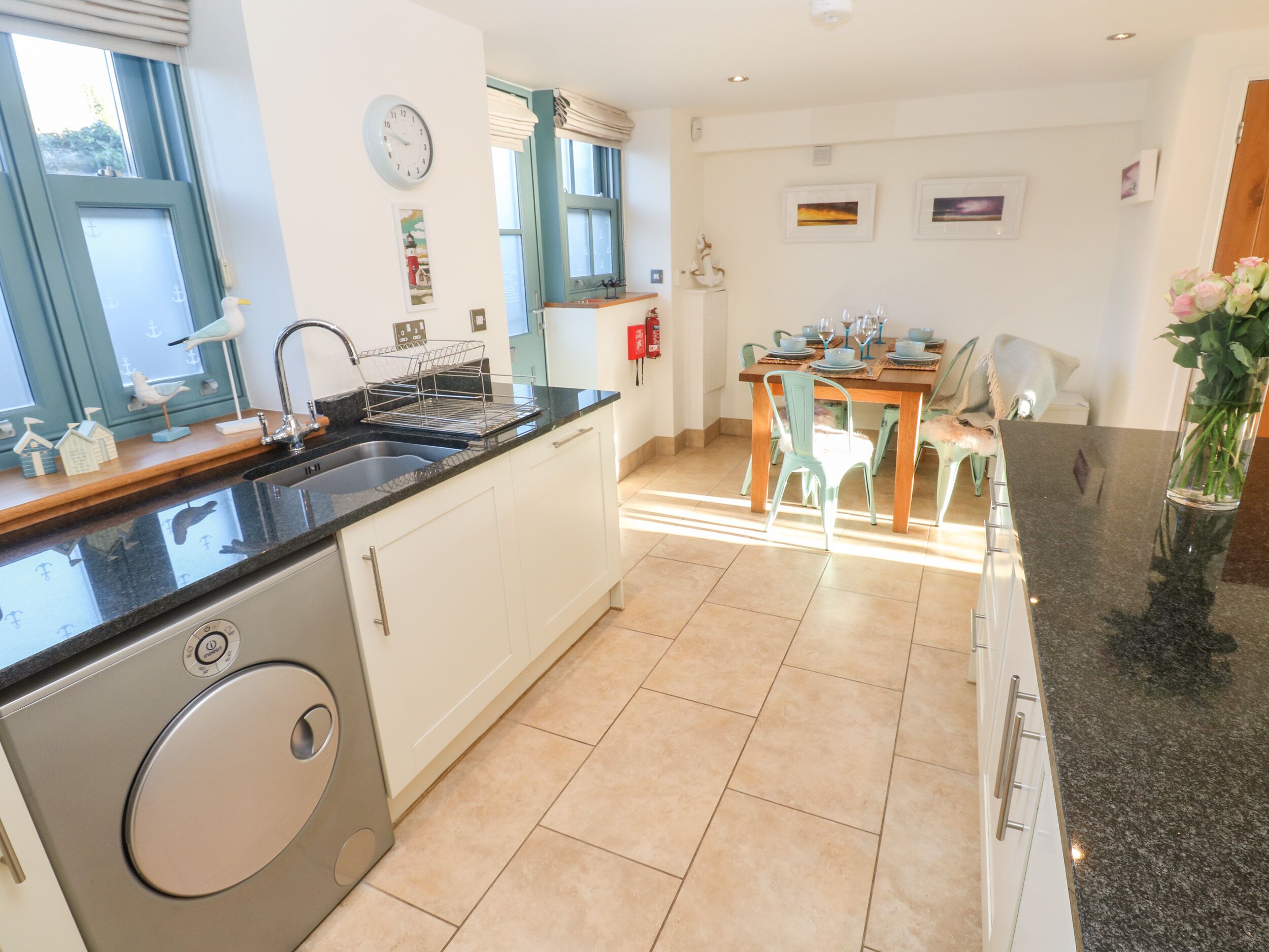 A modern kitchen with a built-in washing machine, a dining area with a wooden table and chairs, and a view of the garden through the window.