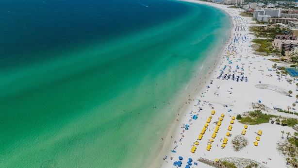 Vue aérienne d’une plage aux eaux turquoise et limpides, bordée d’un rivage sablonneux et parsemée de nombreux parasols.