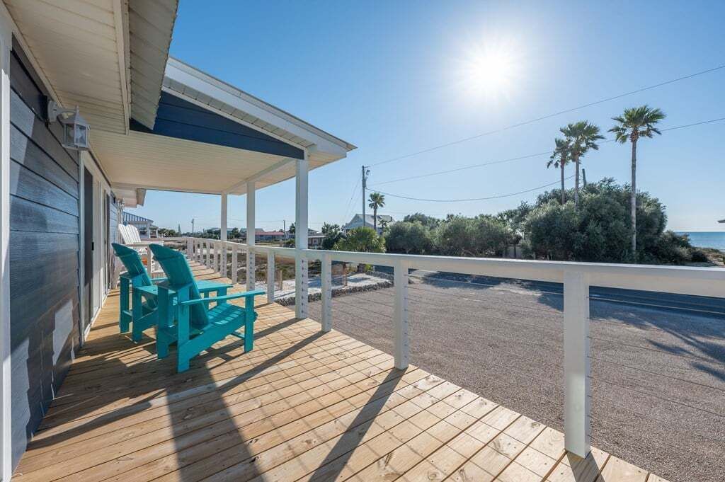 A wooden deck with a covered area, two turquoise chairs, and a view of the beach and palm trees.