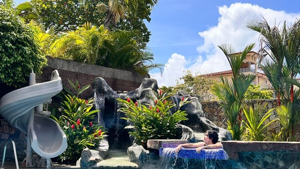 A pool with a water slide, a rock formation, and a person relaxing on a tiled ledge. - Huge Private Home - Exclusive Beach Access and Breathtaking Rooftop Sunset Views (Jaco beach)