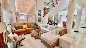 A spacious living area with a sofa, a coffee table, and a wicker chair. A staircase leads to an upper level. - Huge Private Home - Exclusive Beach Access and Breathtaking Rooftop Sunset Views (Jaco beach)