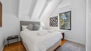 Ein modernes Schlafzimmer mit schrägverdachter Decke, einem großen Bett mit weißer Bettwäsche, einem Nachttisch mit Lampe und einem Fenster mit Blick auf Bäume.