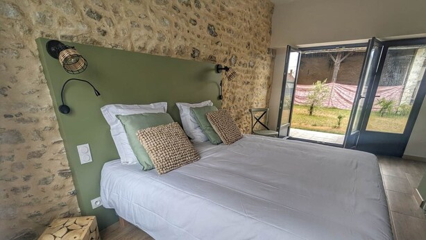 A bedroom with a stone wall, a large bed with white bedding, two wall-mounted lamps, and a view of a garden through a glass door.