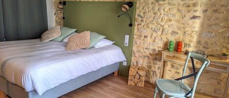 Quarto com parede de pedra, cama com cabeceira cinza, mesa de cabeceira de madeira e uma cadeira.