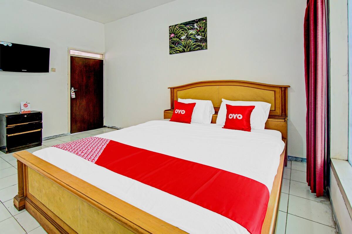 A neatly made bed with a red and white bedspread, two red pillows with "OYO" branding, a wooden headboard, a television mounted on the wall, and a door in the background.