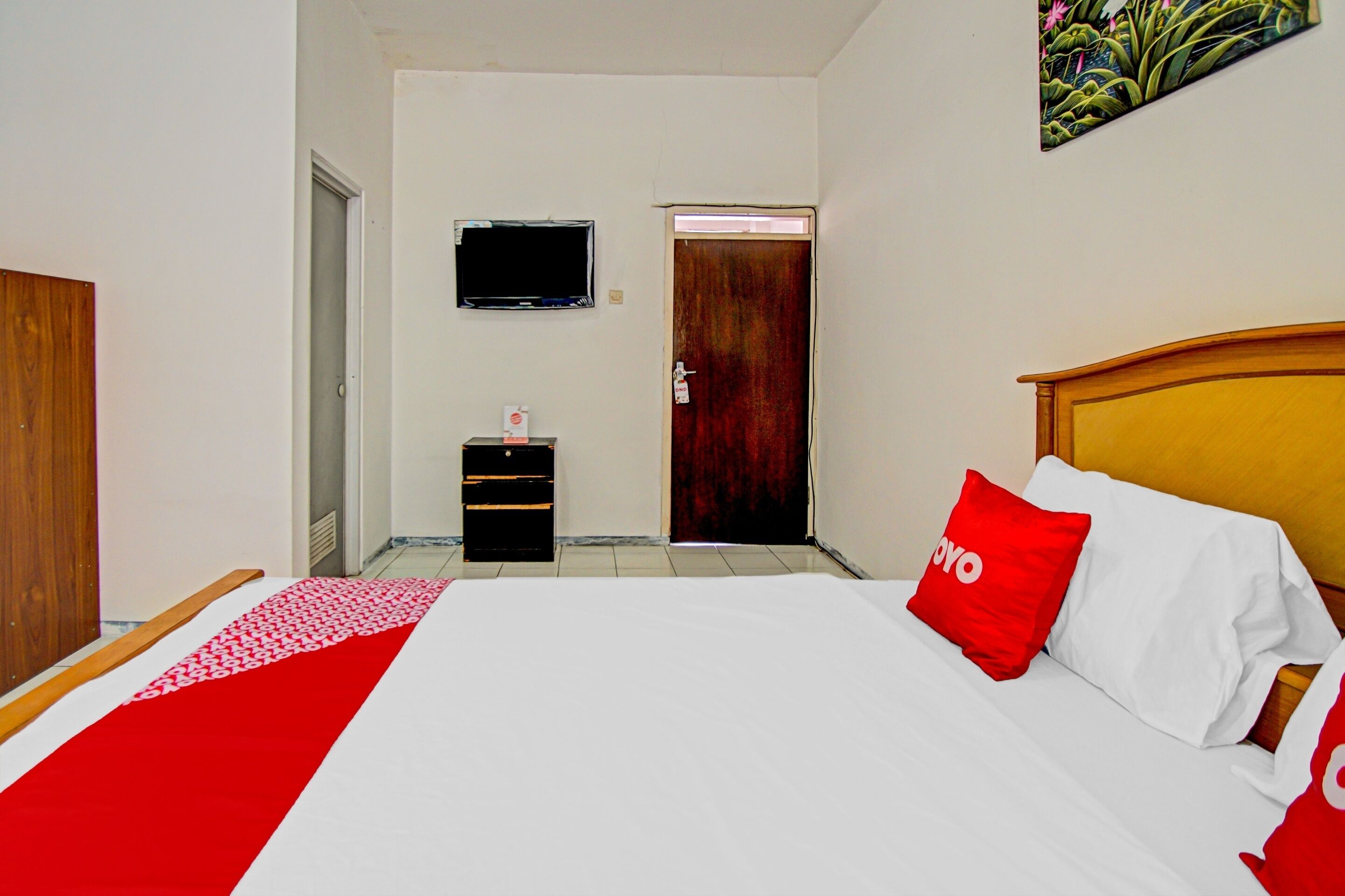 A hotel room with a bed, a wooden headboard, a red and white bedspread, a red pillow with the word "YOYO", a television mounted on the wall, and a door leading to another room.