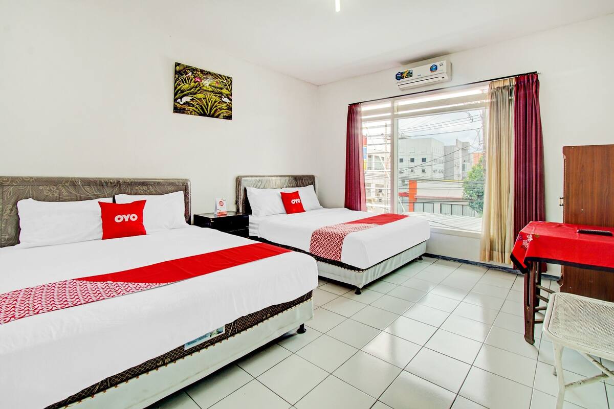A hotel room with two beds, a desk, a chair, a window with curtains, an air conditioner, and a painting on the wall.
