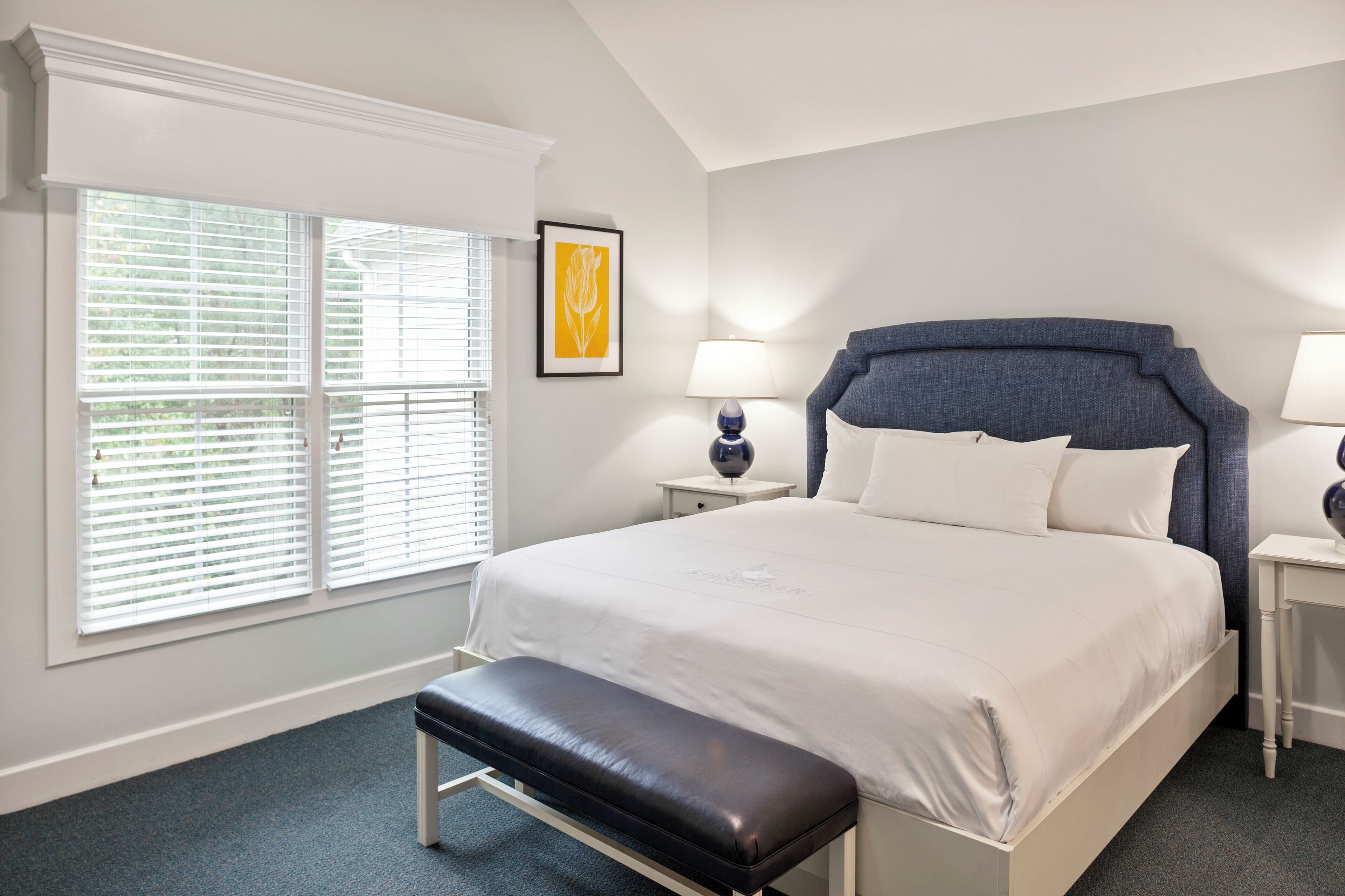 A neatly arranged bedroom with a large bed, a bench, two bedside tables, and a window with blinds.
