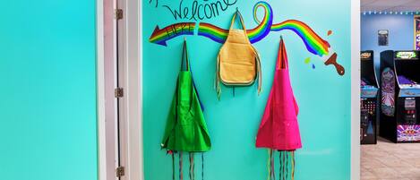 En teal-farvet væg med teksten "Artists Welcome" og et regnbue-design, samt tre tasker hængende på kroge.