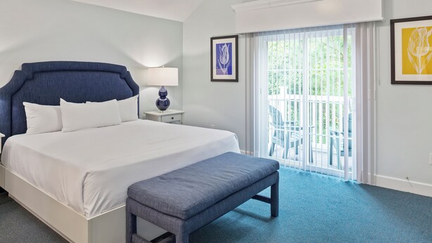 A hotel room with a large bed, a bench, a nightstand, and a view of the outdoors through a sliding glass door. - King’s Creek Resort by Spinnaker (Williamsburg)