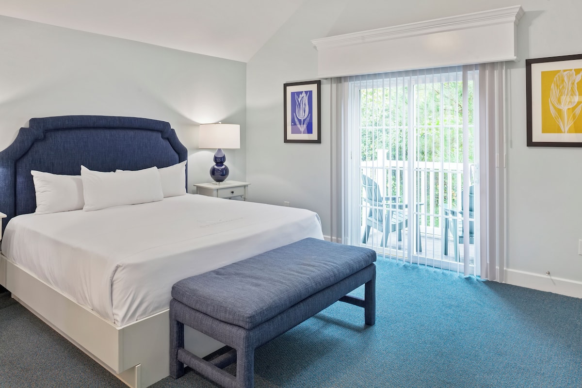 A hotel room with a large bed, a bench, a nightstand, and a view of the outdoors through a sliding glass door.