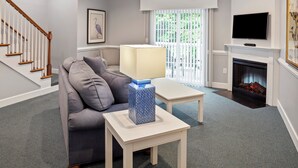 Un salón con un sofá gris, una mesa de centro blanca con un jarrón azul, una chimenea y un televisor montado encima.