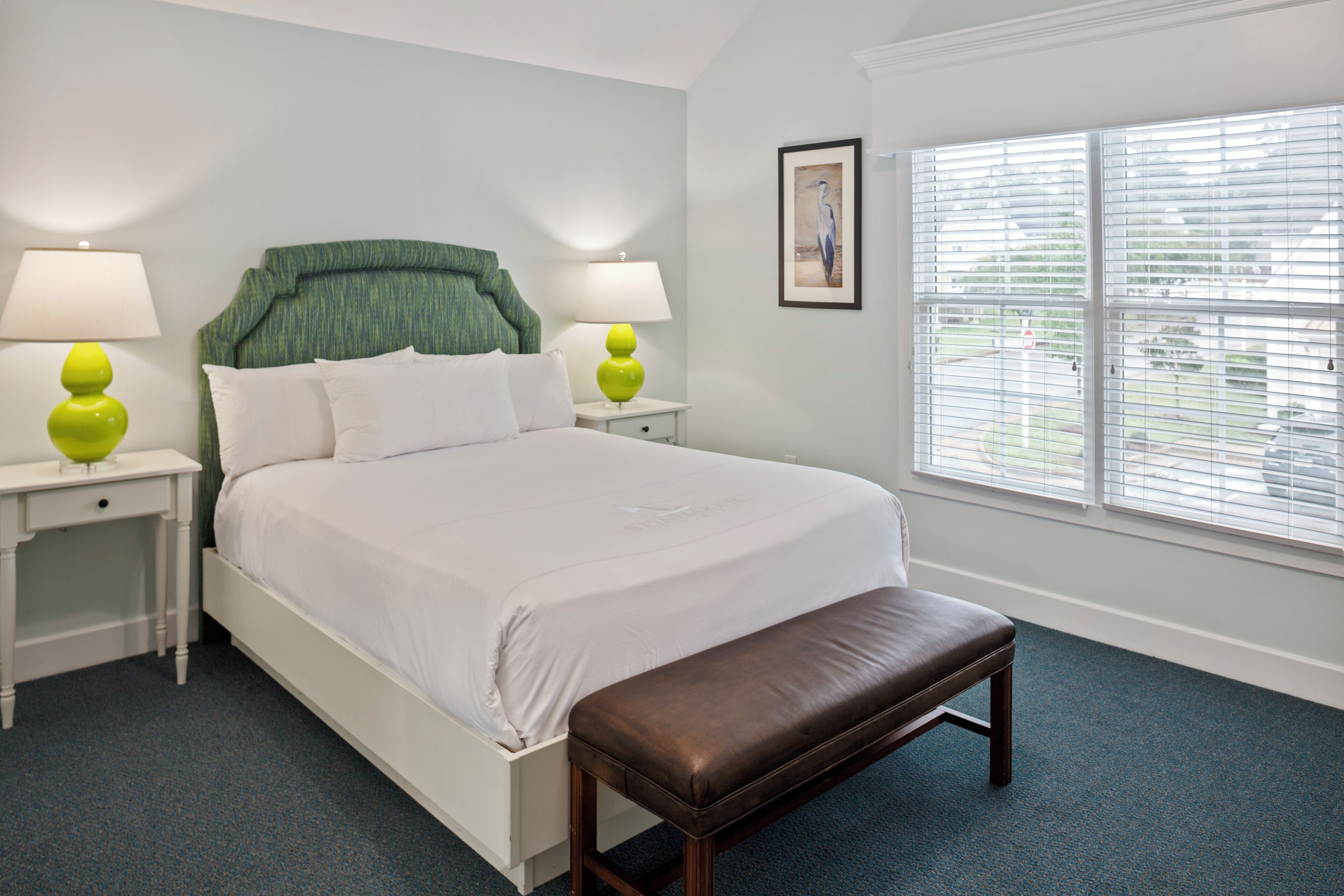A bedroom with a bed, two bedside tables, a bench, a window with blinds, and a framed picture on the wall.