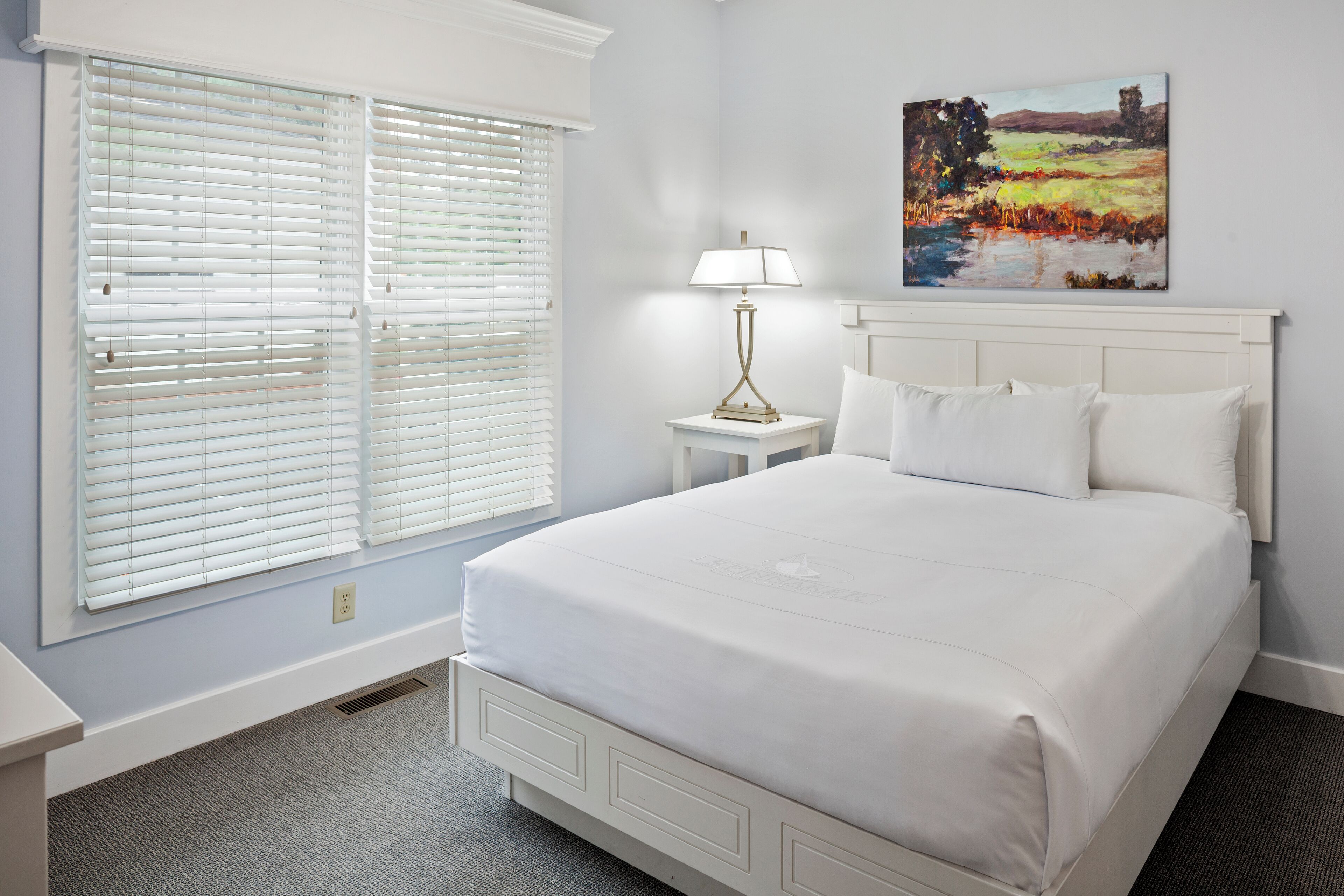 A bedroom with a large bed, white bedding, a nightstand with a lamp, and a painting on the wall.