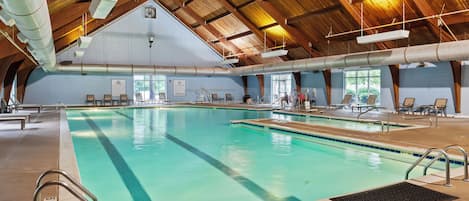 An indoor swimming pool with a high ceiling and wooden beams, surrounded by lounge chairs and a clear view of the outdoors.