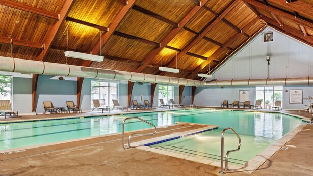 Una piscina interior con techo de madera, paredes de vidrio transparente y varias sillas de descanso alrededor de la zona de la piscina.
