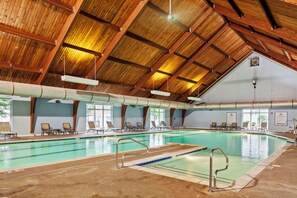 An indoor swimming pool with a wooden ceiling, clear glass walls, and multiple lounge chairs around the pool area. - King’s Creek Resort by Spinnaker (Williamsburg)