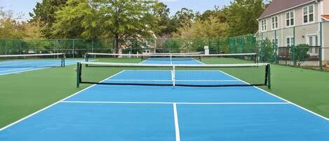 Una cancha de tenis al aire libre con una red en el centro, rodeada por una valla y árboles.