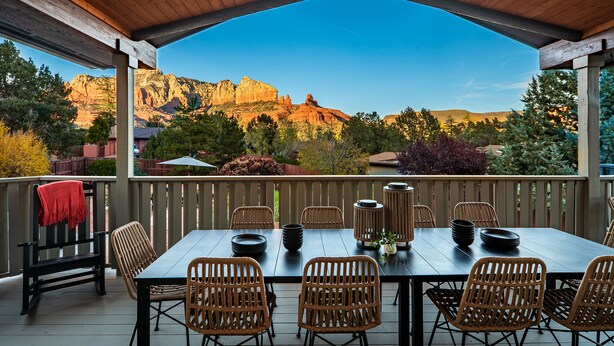 Un patio techado con techo de madera, una mesa dispuesta con platos y un jarrón, sillas de mimbre y vistas a formaciones rocosas rojas.